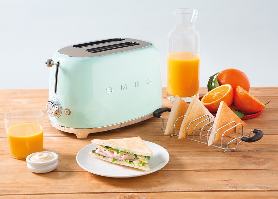 Pastel green two-slot toaster on a wooden table with toasted sandwiches, oranges, and a glass of orange juice
