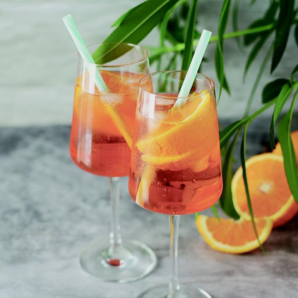 A pair of wine glasses filled with Orange Spritz, ice, straws, and orange slices, set against a textured background with green leaves.