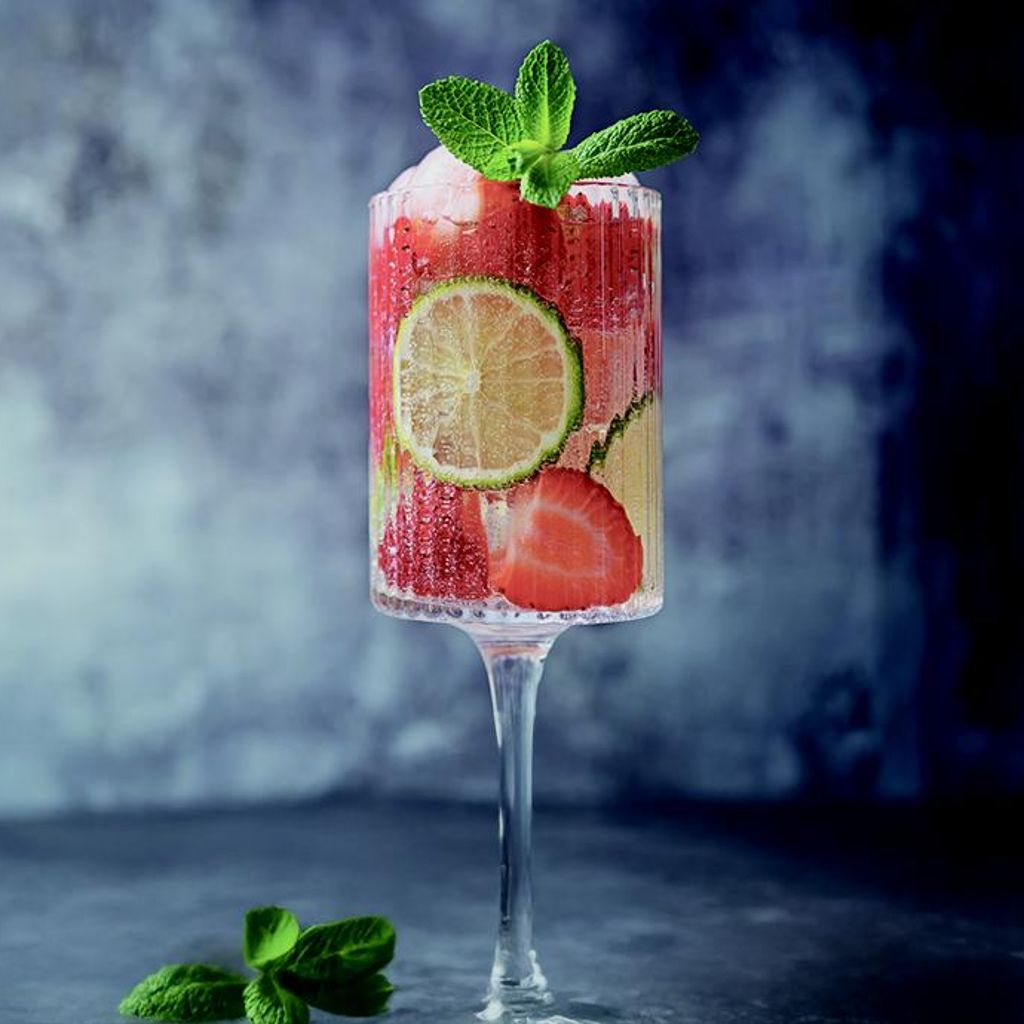 A sparkling drink in a tall glass with lime slices, fresh strawberries, and mint, set against a dark, textured background.