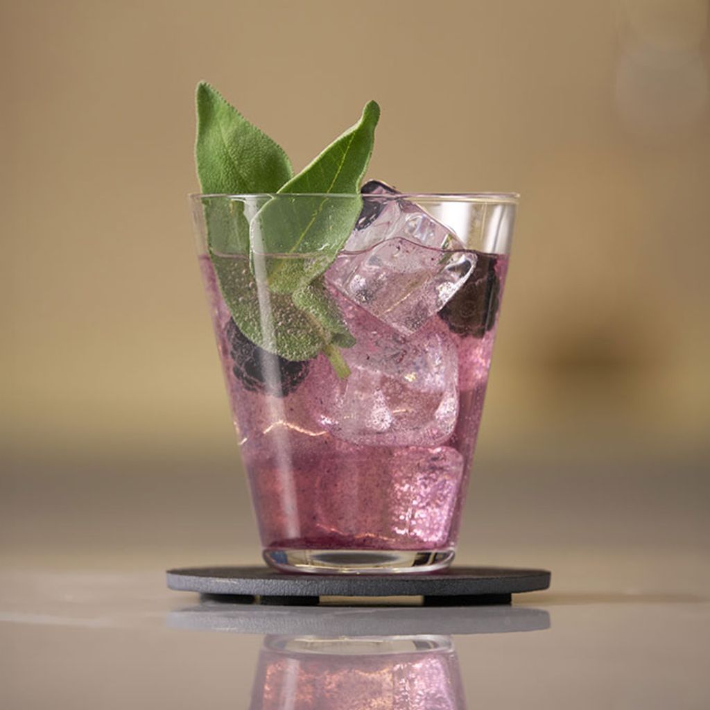 A purple drink with ice, blackberries, and fresh sage leaves in a glass served on a dark coaster.