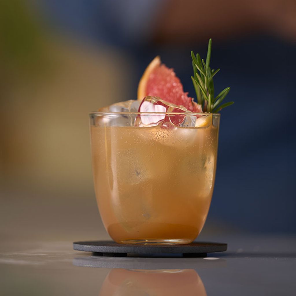 A citrus cocktail served over ice, garnished with a slice of pink grapefruit and a sprig of fresh rosemary.