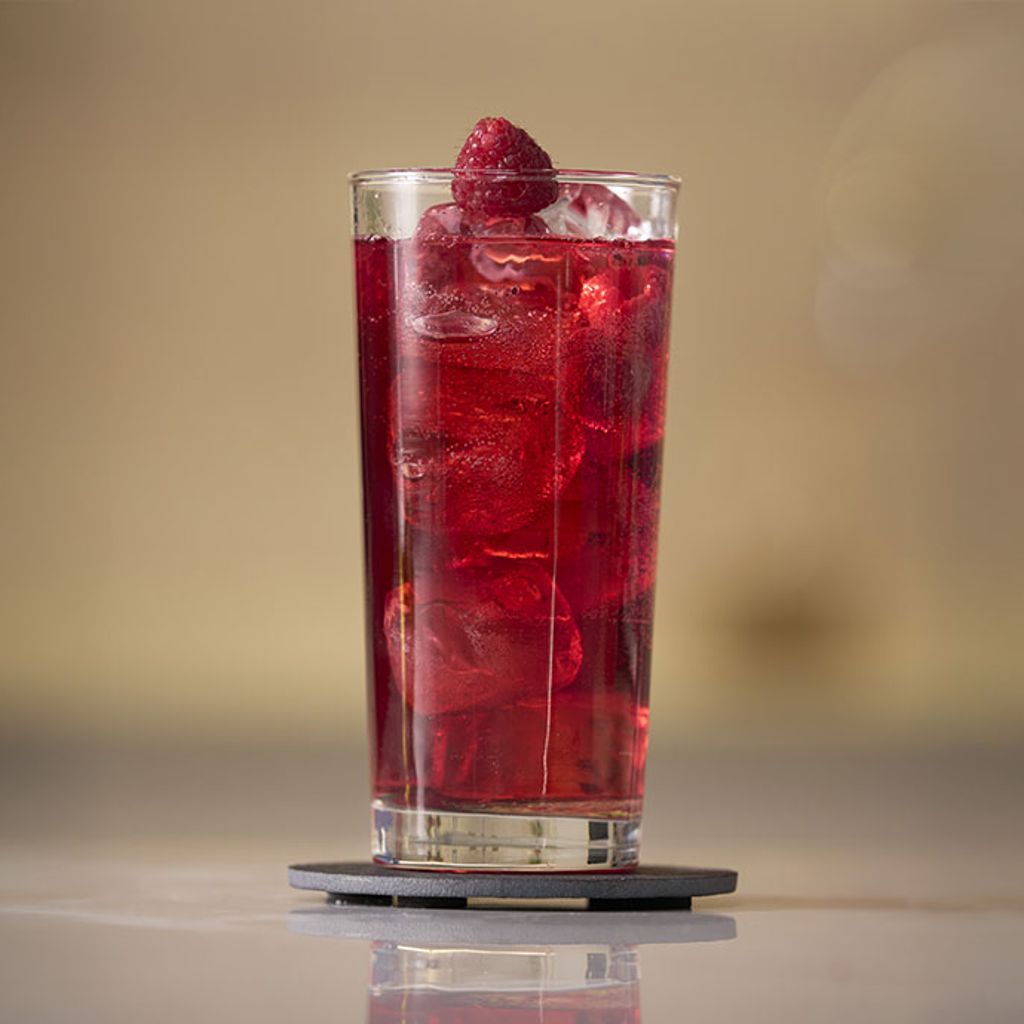 A bright red drink with ice and a raspberry in a tall glass, placed on a dark gray coaster.