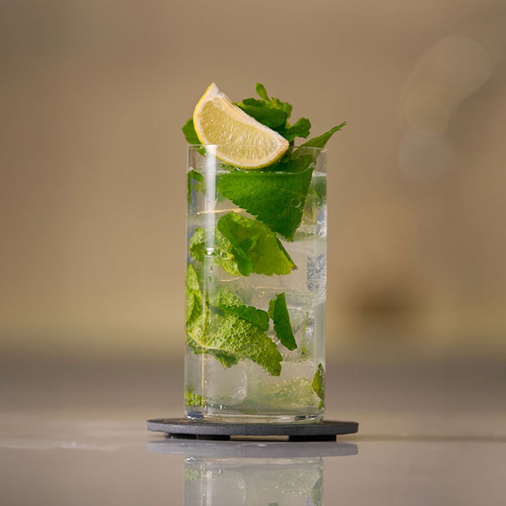 Un bicchiere alto riempito con un cocktail trasparente, guarnito con foglie di menta fresca e ghiaccio, e decorato con una fetta di lime giallo.