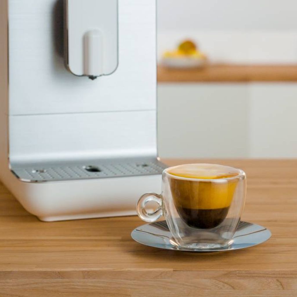 Soy and turmeric cappuccino served in a clear glass cup on a wooden countertop next to a modern coffee machine, with layered espresso and golden foam visible.