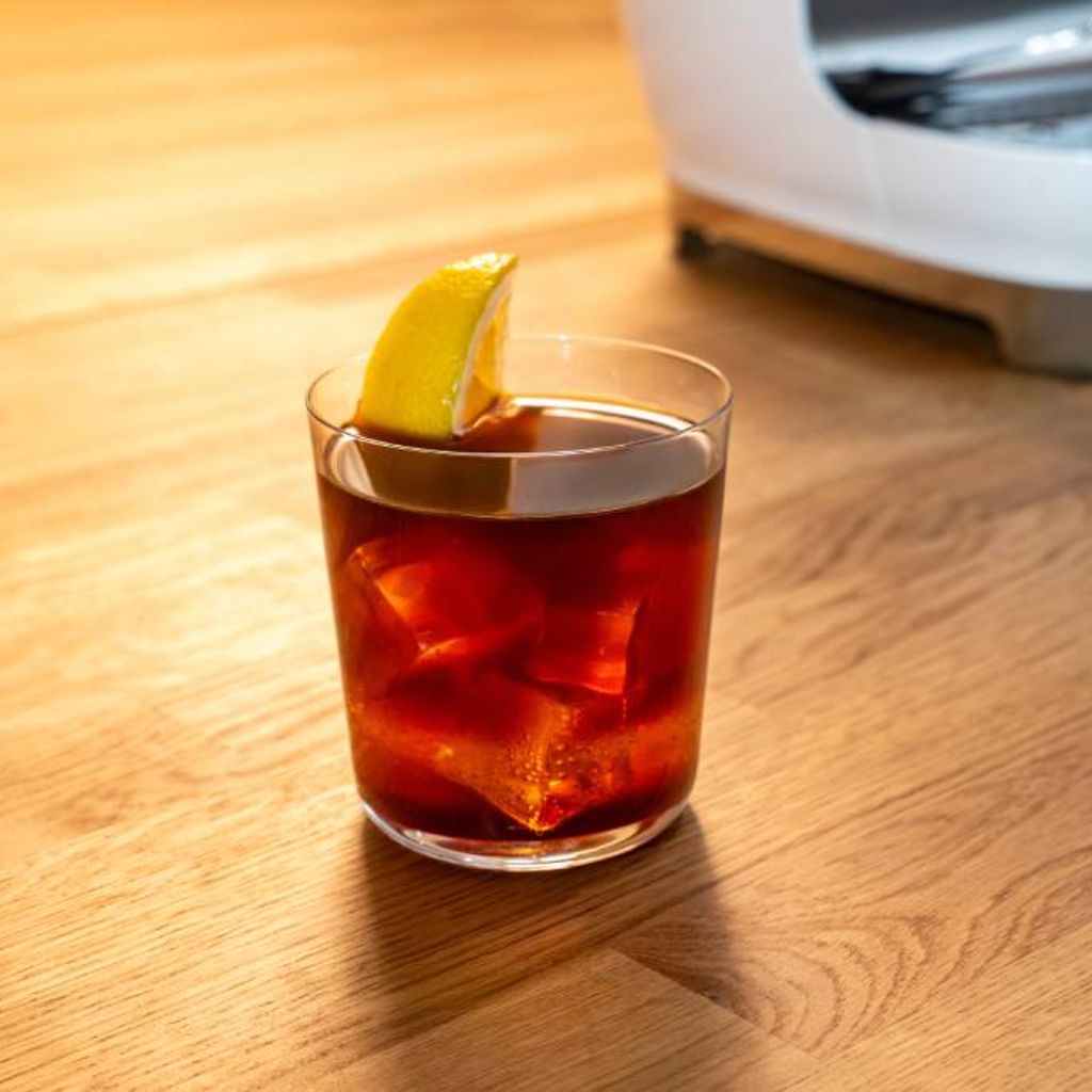 Glass of cold brew coffee with ice and a slice of lemon on a wooden table, with a white SMEG coffee machine.