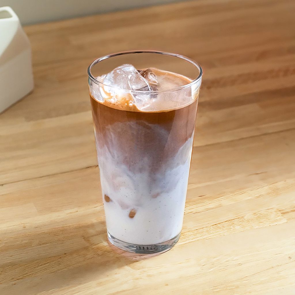 A tall glass with layers of milk and coffee and ice, on a wooden table next to a small white vase.