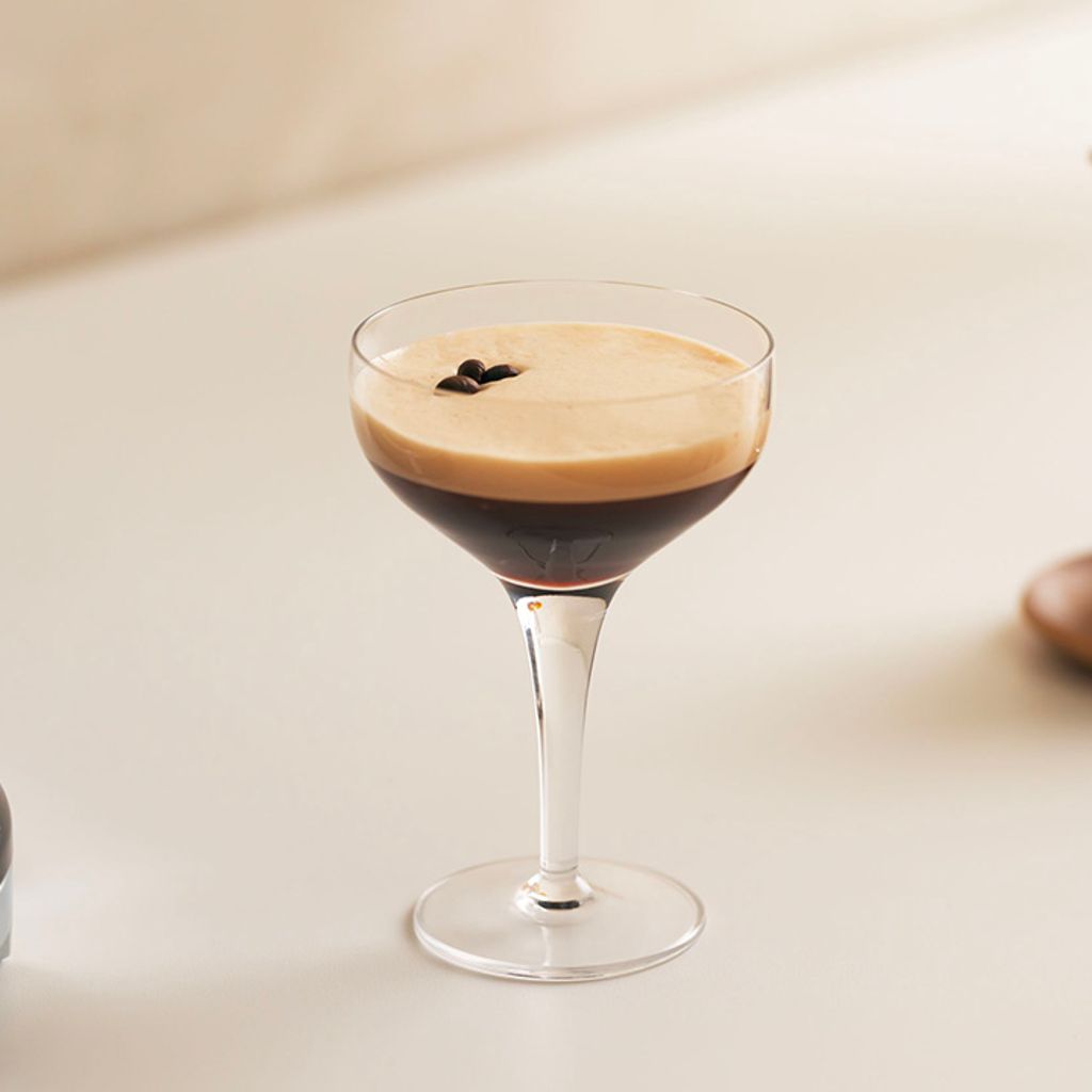 A cup of two-layered shaken coffee with thick foam, garnished with coffee beans, next to a black Smeg coffee machine.