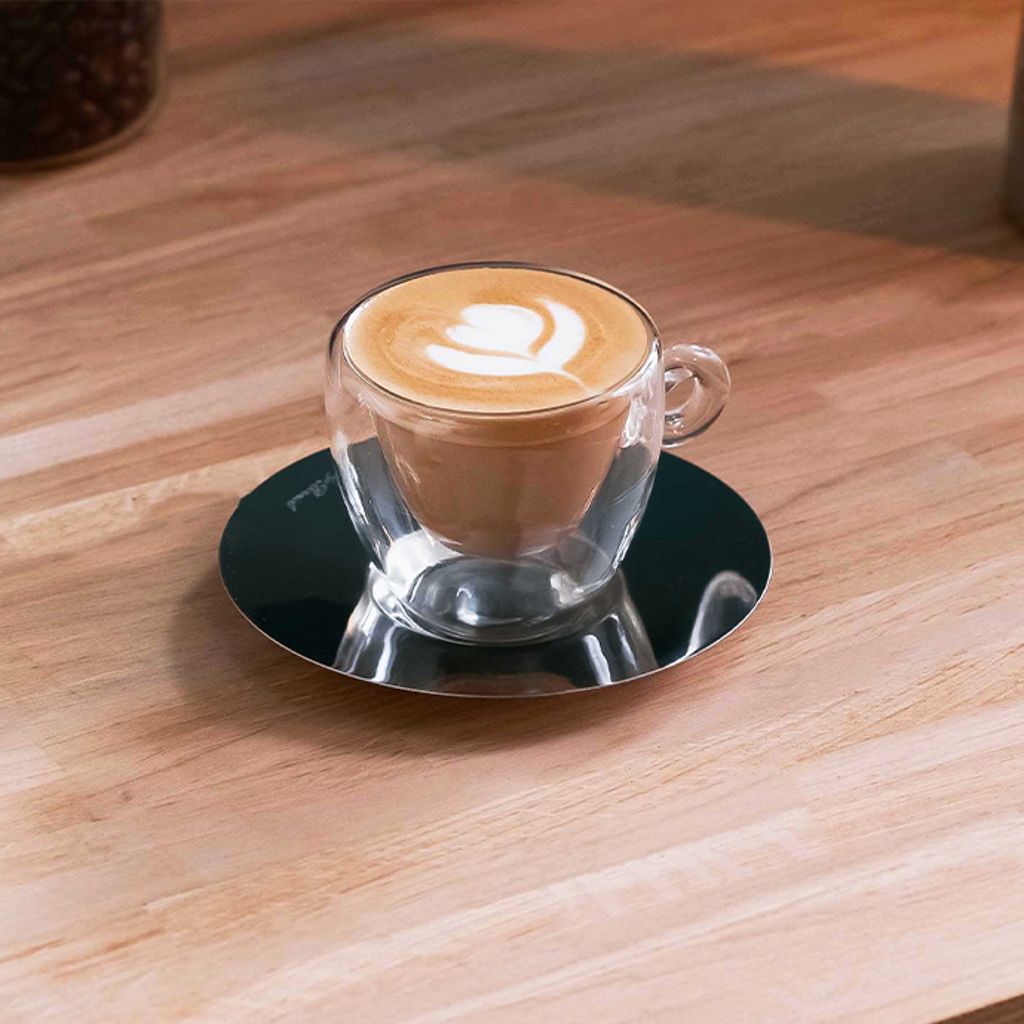 Cappuccino with latte art in a transparent glass cup on a dark reflective saucer, placed on a wooden table.