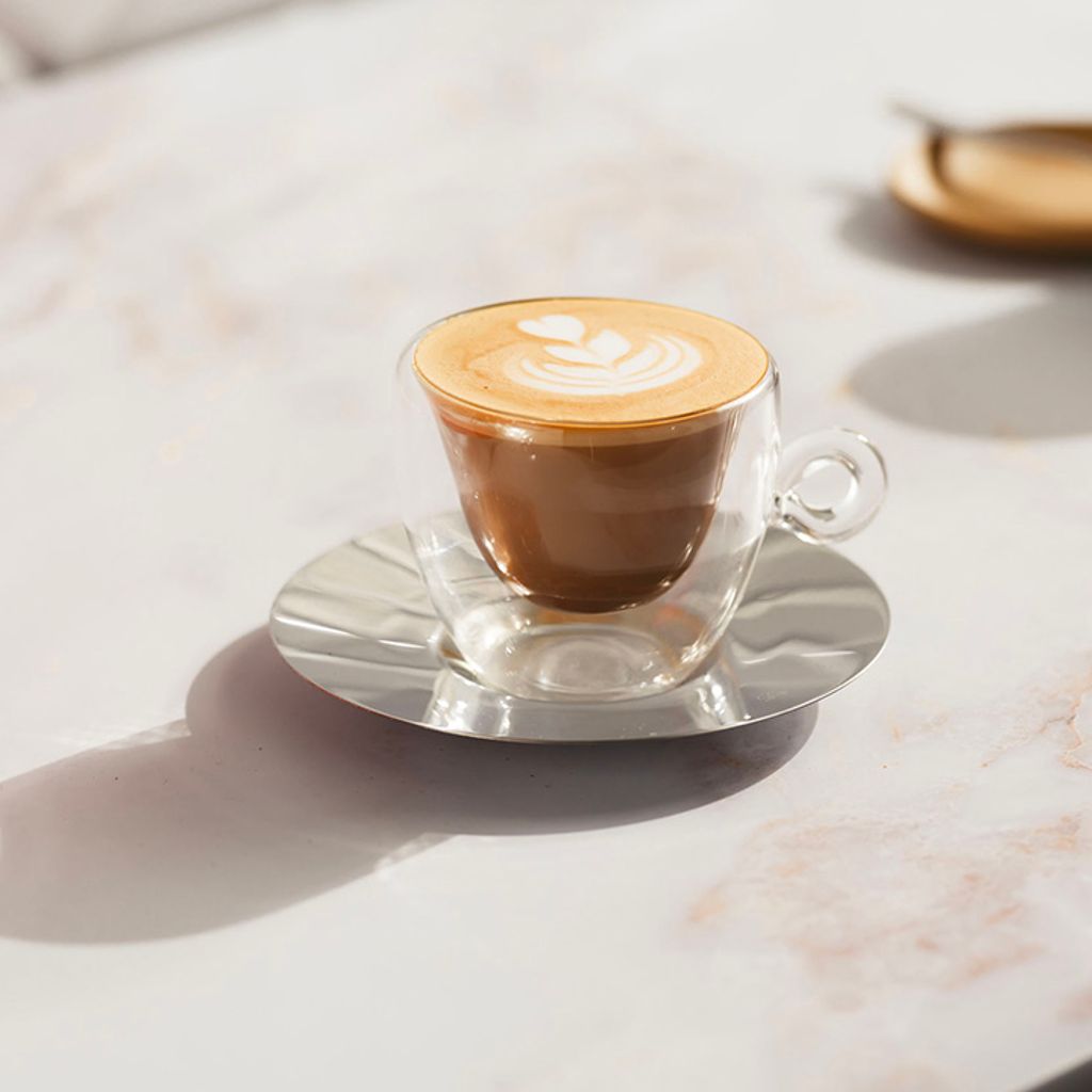 Transparent glass cup with coffee and decorated foam, on a steel saucer atop a light marble countertop.
