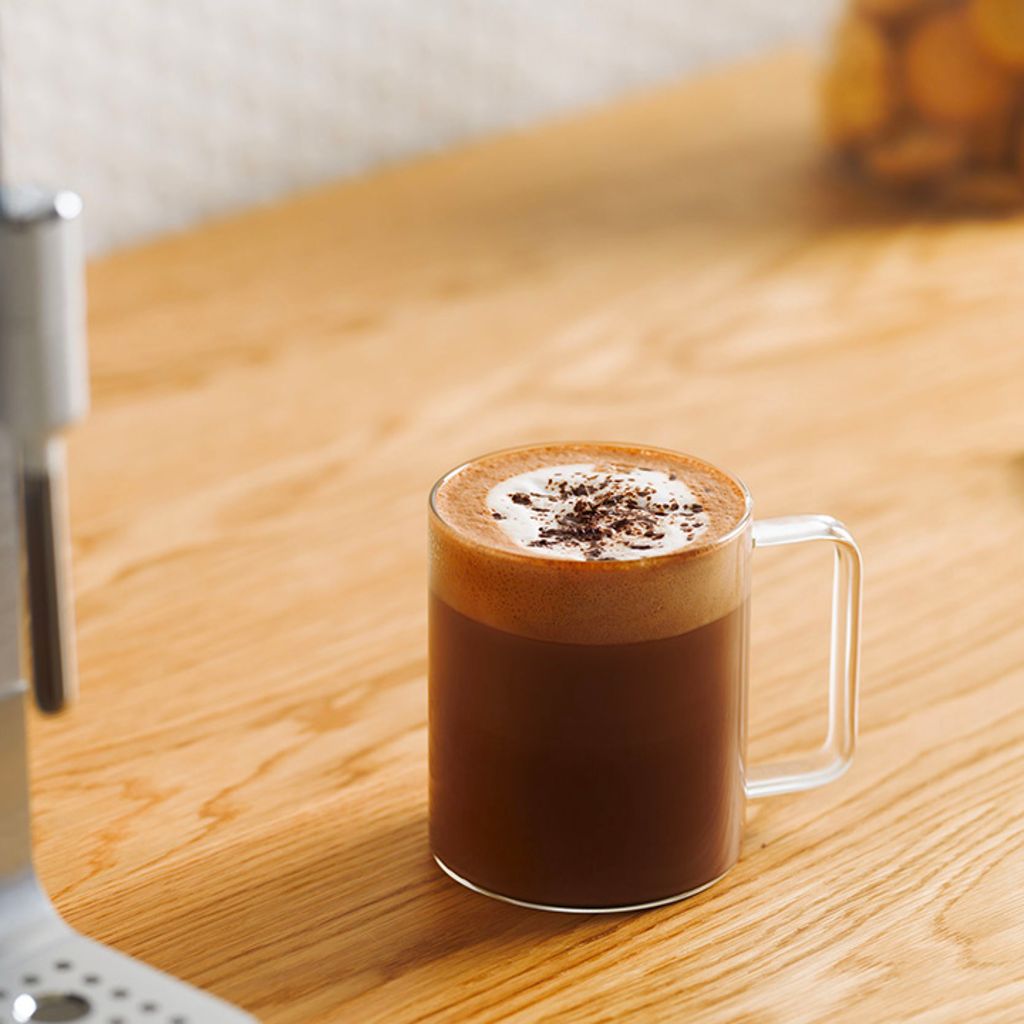 A glass cup of mocha with a layer of white foam and chocolate shavings on a wooden countertop next to a Smeg coffee machine.
