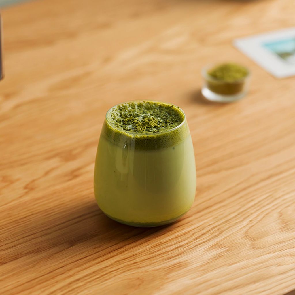 A rounded glass containing matcha latte with thick foam dusted with green powder, on a light-colored wooden table.