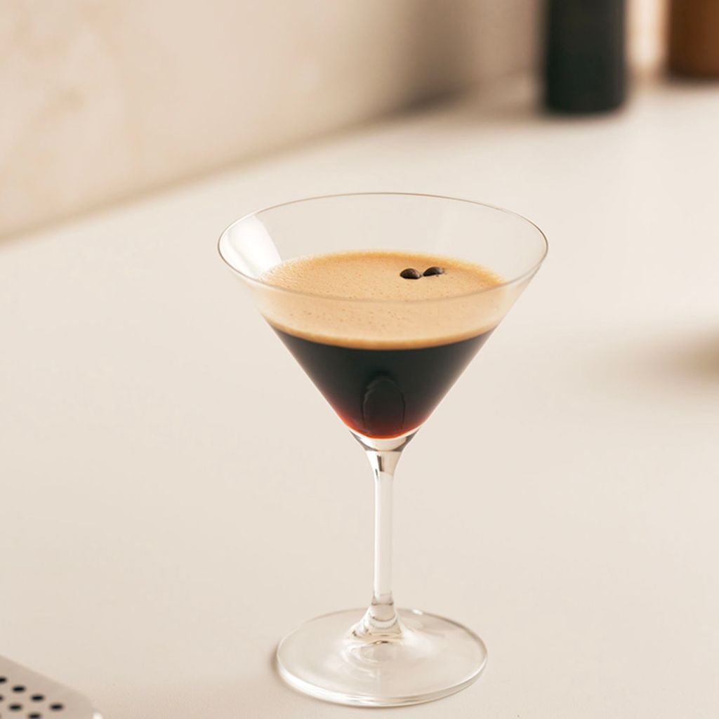 An Espresso Martini with thick foam and coffee beans in a crystal glass, next to a Smeg coffee machine.