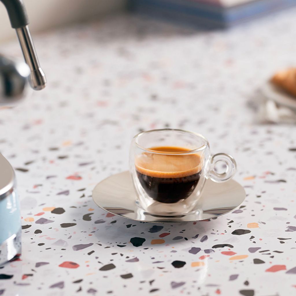 Espresso with a golden crema in a clear cup and stainless steel saucer, next to a light blue Smeg coffee machine.