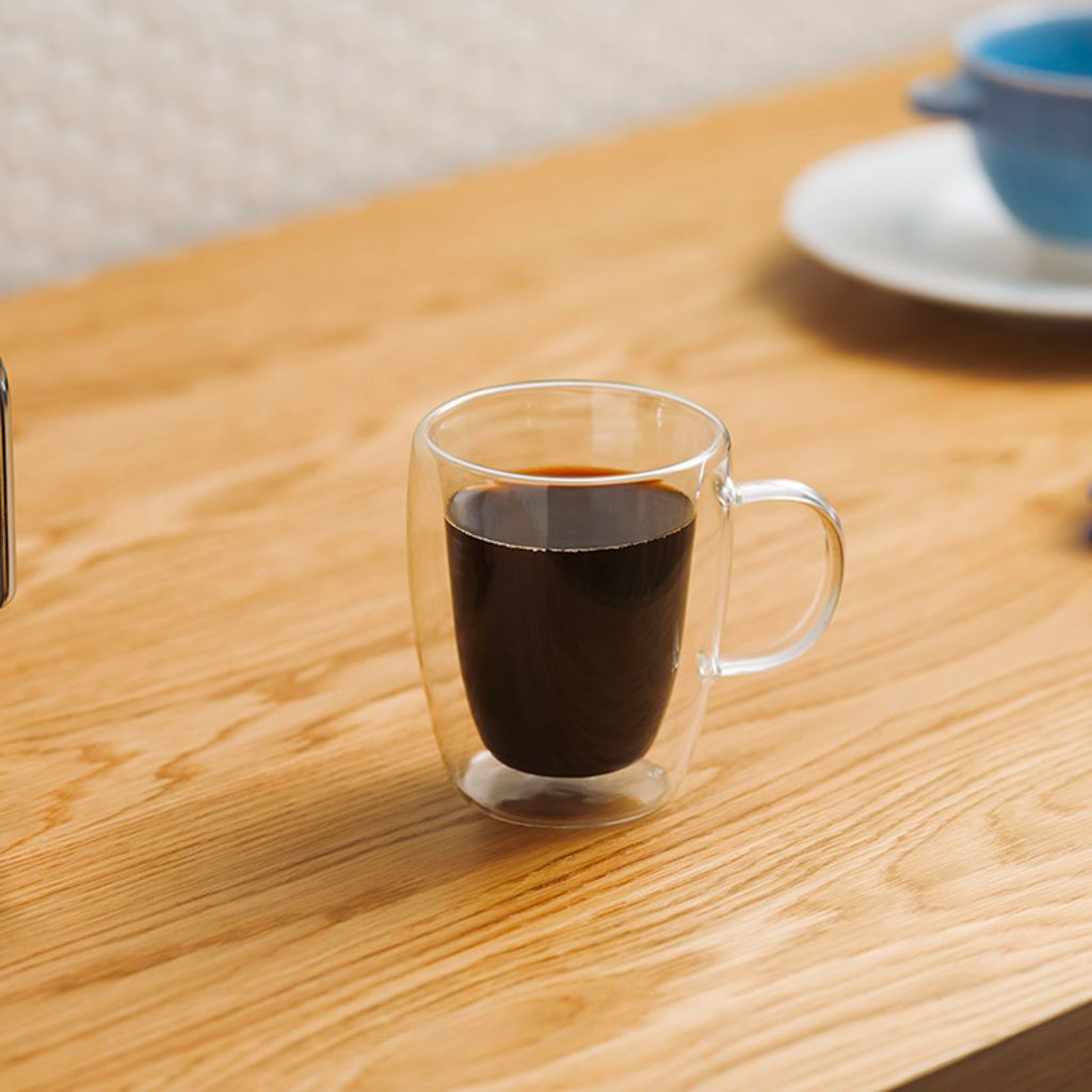 Una tazza di vetro trasparente con del caffè nero accanto a una macchina da caffè a filtro Smeg su un tavolo di legno chiaro.