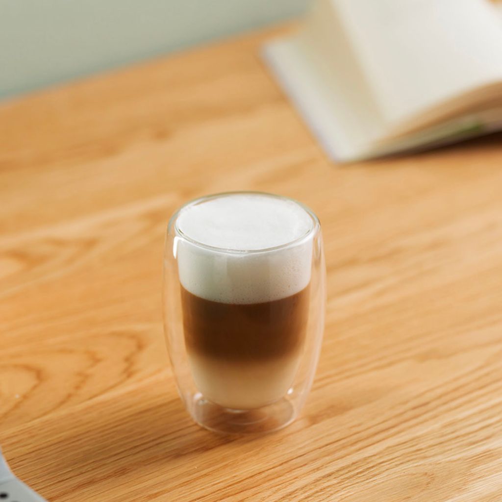 A tall glass with distinct layers of milk, coffee, and foam, sitting on a wooden table against a blurred background.