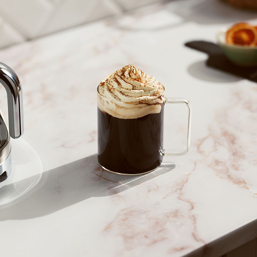 Ginger coffee in a glass cup topped with a generous dollop of whipped cream, served on a marbled countertop next to a Smeg coffee machine.