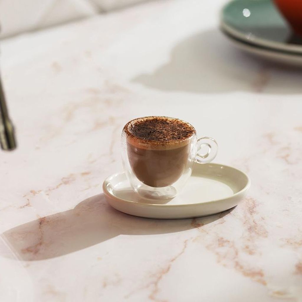 Marocchino coffee with foam and cocoa in a clear cup on a pink marble countertop, next to a red Smeg coffee machine.