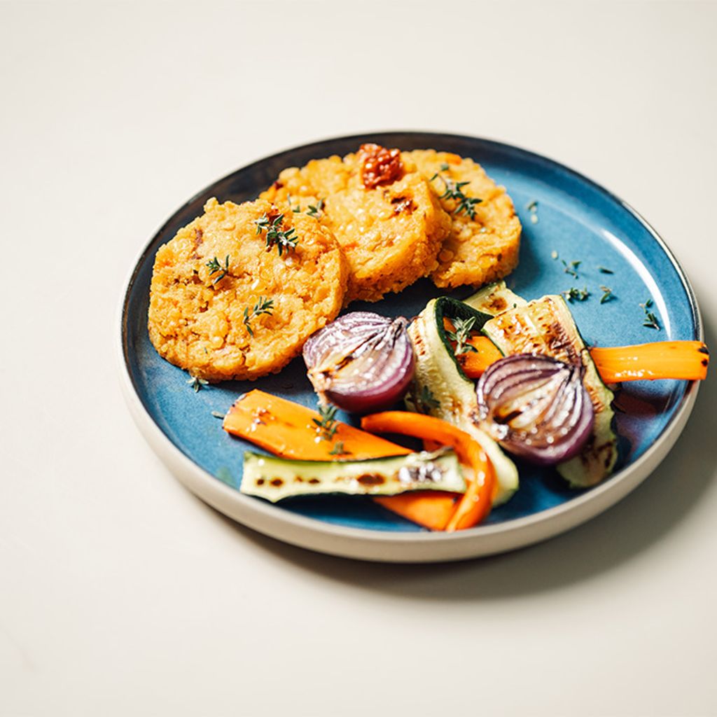 A blue ceramic plate with three golden lentil patties garnished with herbs, served alongside grilled vegetables including sliced zucchini, red onion halves, and carrot strips on a light-colored surface.