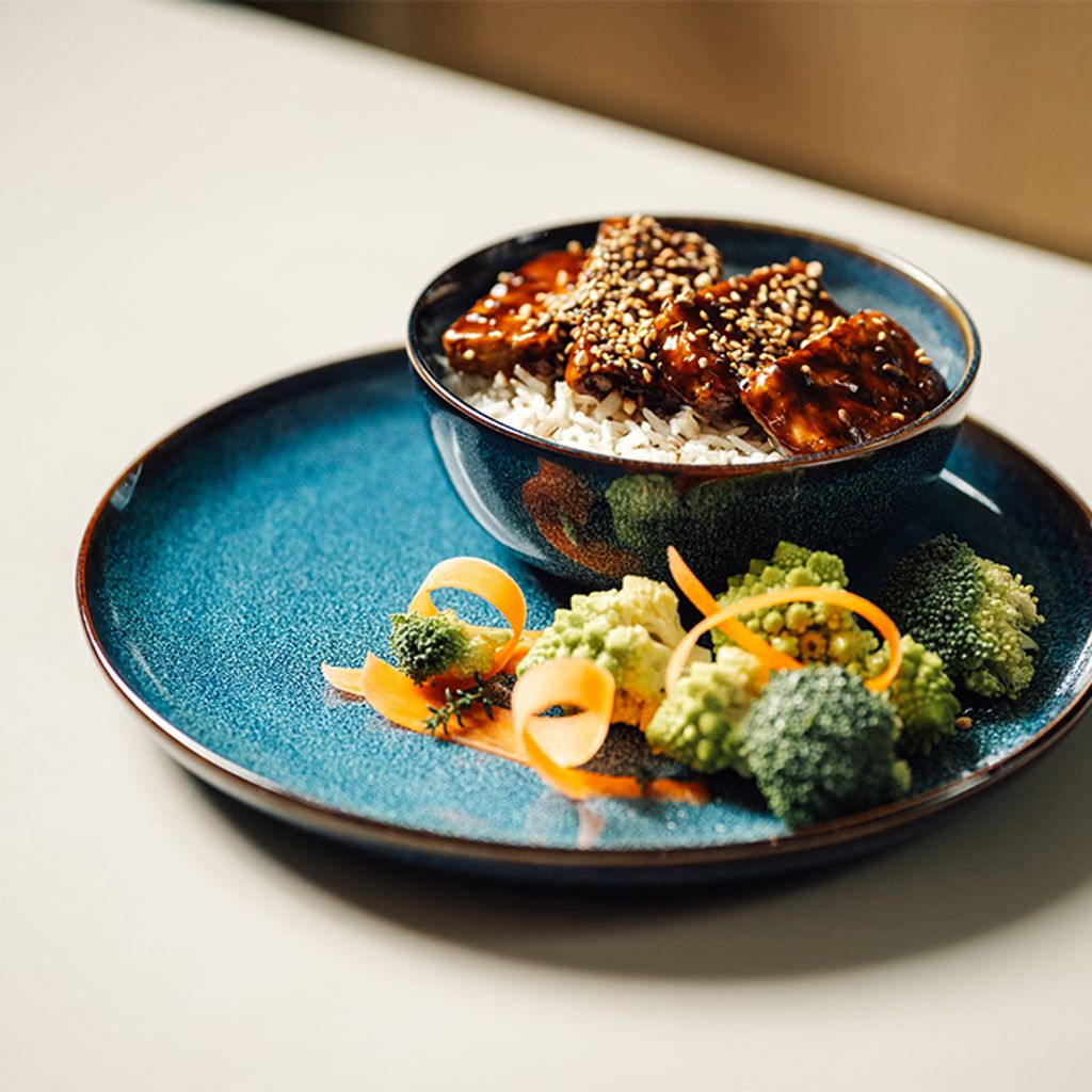 Blue bowl with white rice and slices of tofu glazed with teriyaki sauce and sesame seeds, served with broccoli and spiralized carrots.