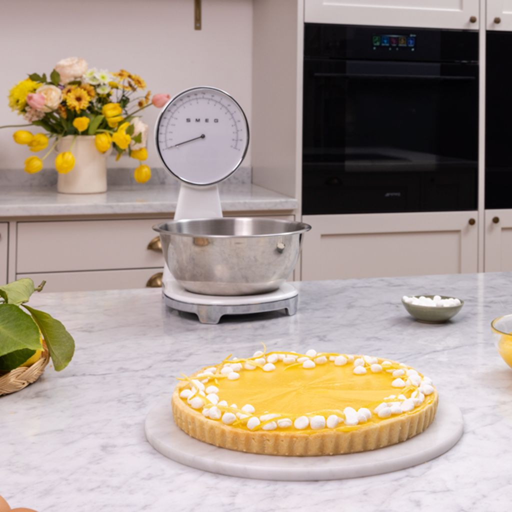 Lemon tart with white meringue on a marble countertop, featuring a Smeg scale, fresh lemons, and ovens in the background.
