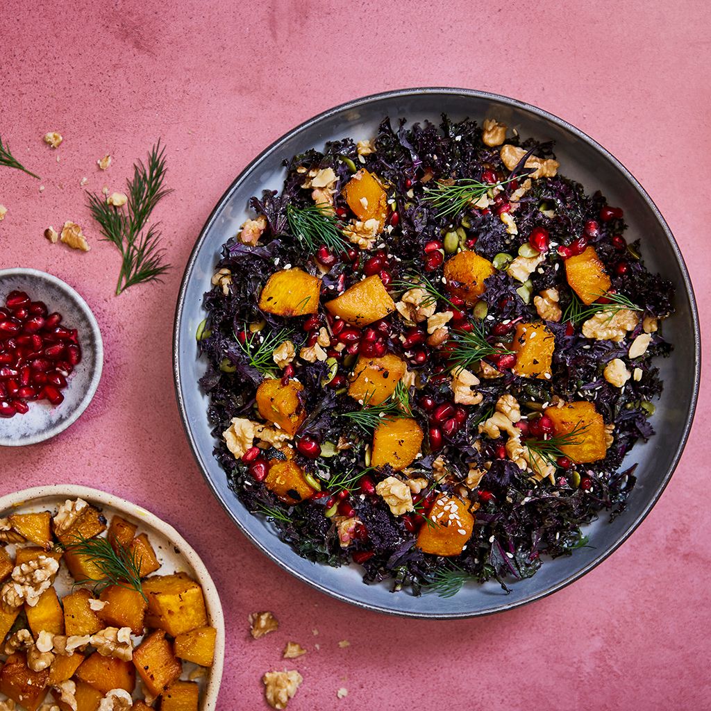 Dark kale salad with slices of orange pumpkin, pomegranate seeds and walnuts in a brown bowl.