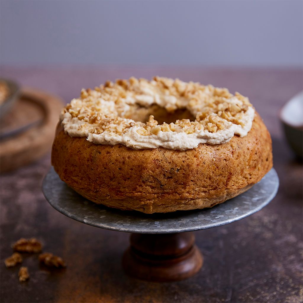 Ciambella di carote con glassa bianca e granella di noci su alzatina in metallo, con sfondo sfocato di noci e ciotole.