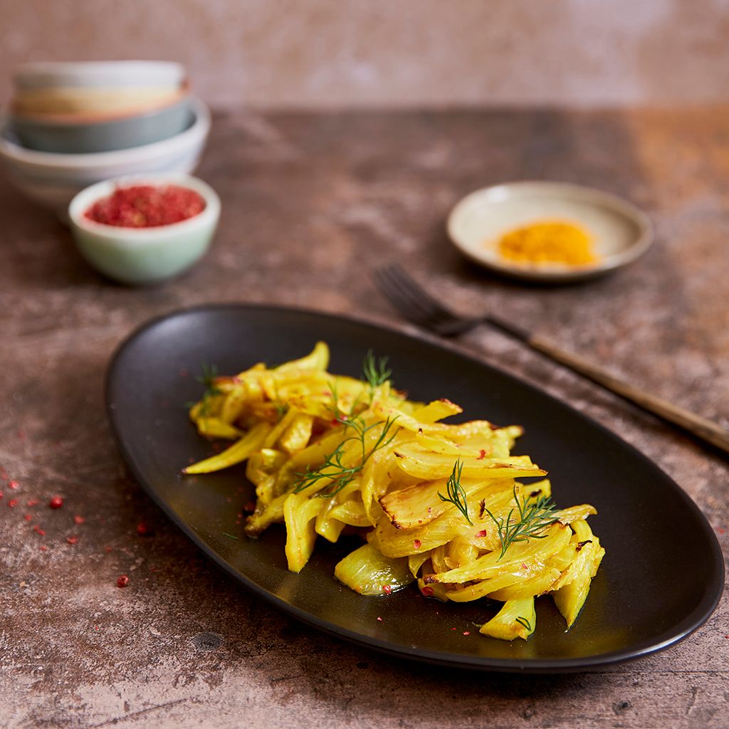 Yellow fennel with turmeric, garnished with dill, served on a black oval plate, with pink berries and small bowls in the background.