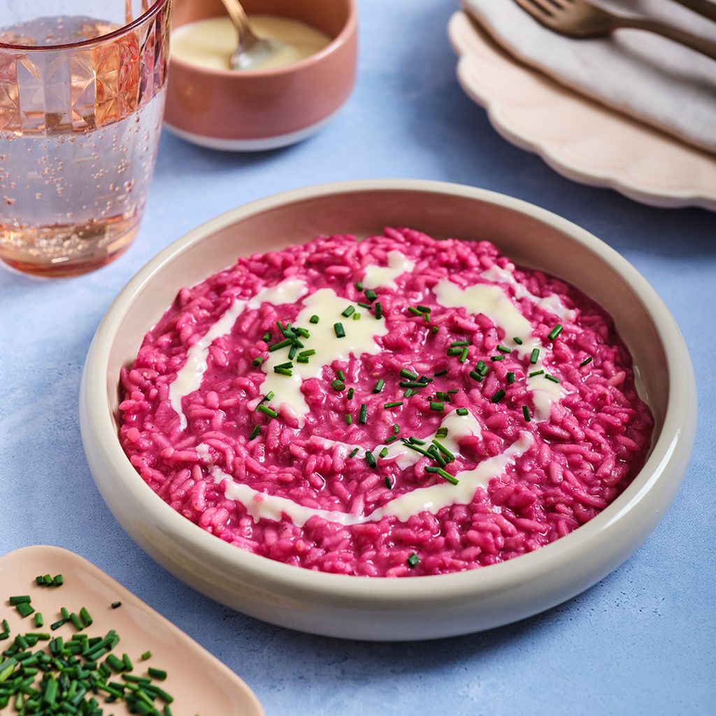 Bright fuchsia risotto in a light-colored bowl, topped with a spiral of white sauce and chives, on a blue background.
