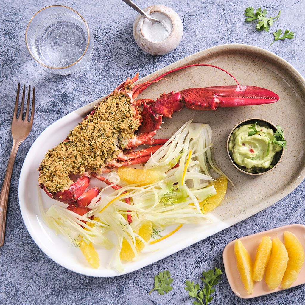 A halved lobster gratin served on a platter with sliced fennel, orange segments, and a side of guacamole.