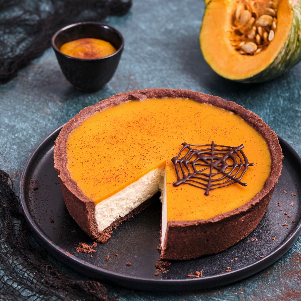 A pumpkin cheesecake with a dark crust, orange topping, and a chocolate spider web, shown with a slice removed.
