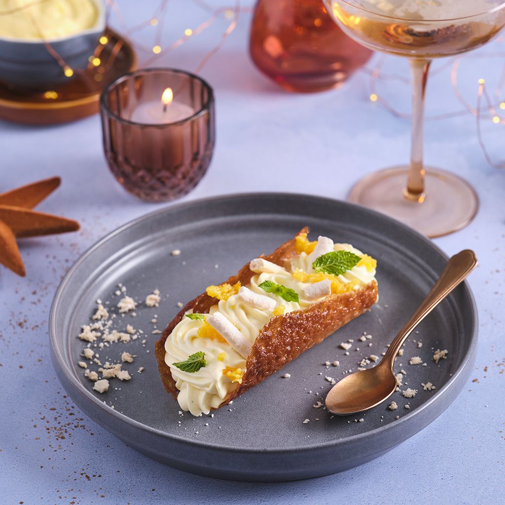A crispy wafer filled with cream, mint and orange on a grey plate, with a candle and a wine glass in the background.