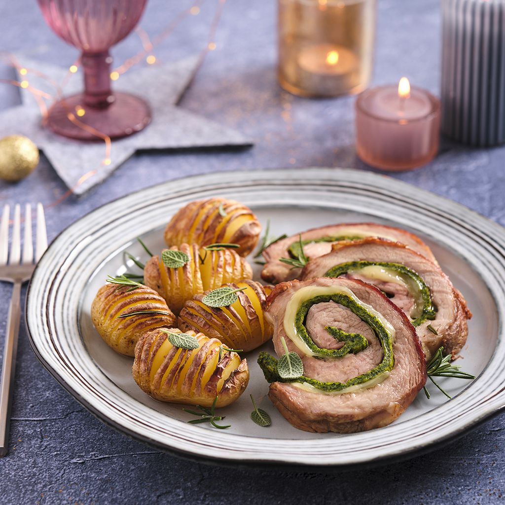 Slices of veal roulade with green stuffing and golden Hasselback potatoes on a flat plate against a Christmas backdrop.