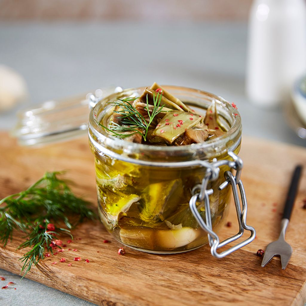 A glass jar of artichokes in oil, with dill and pink peppercorns, on a wooden chopping board with garlic cloves.
