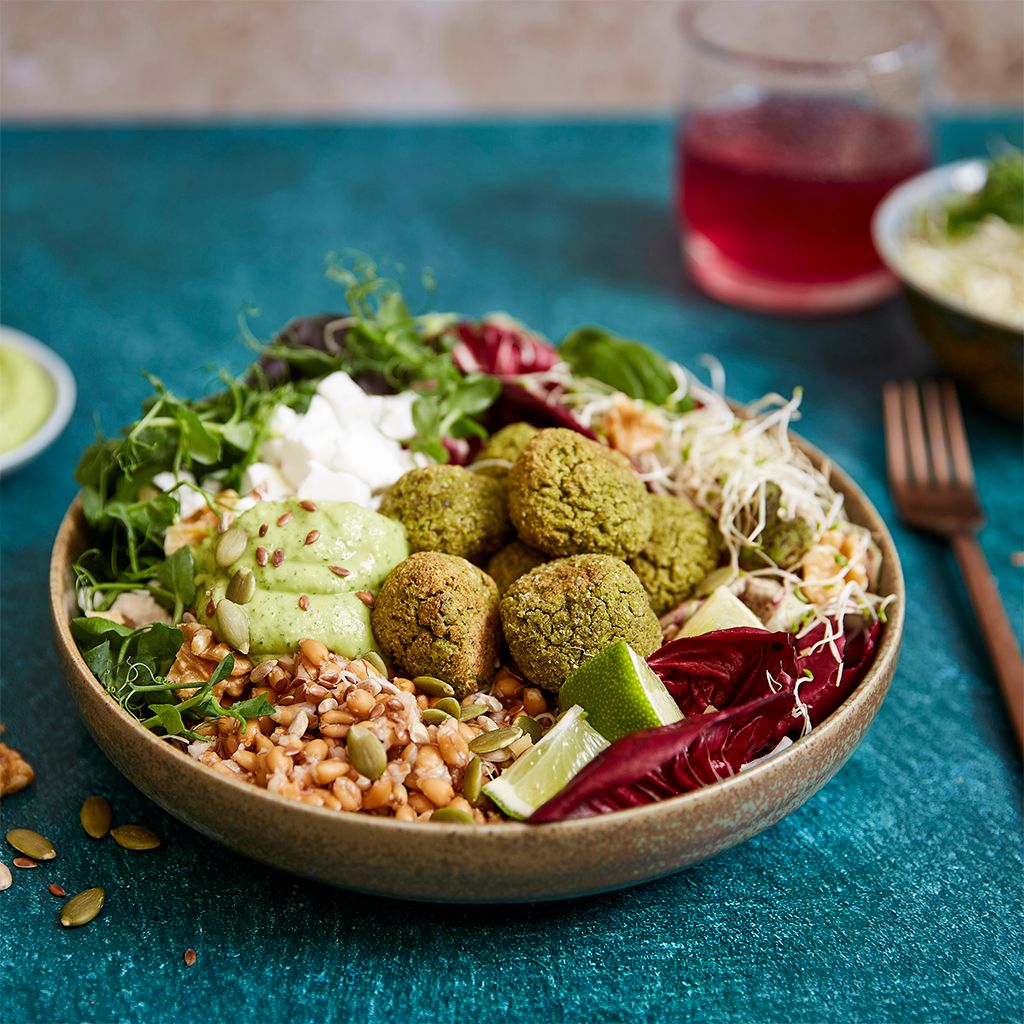 A bowl containing green meatballs, grains, rocket, radicchio and sauces against a light blue background, with seeds and nuts scattered over the top.