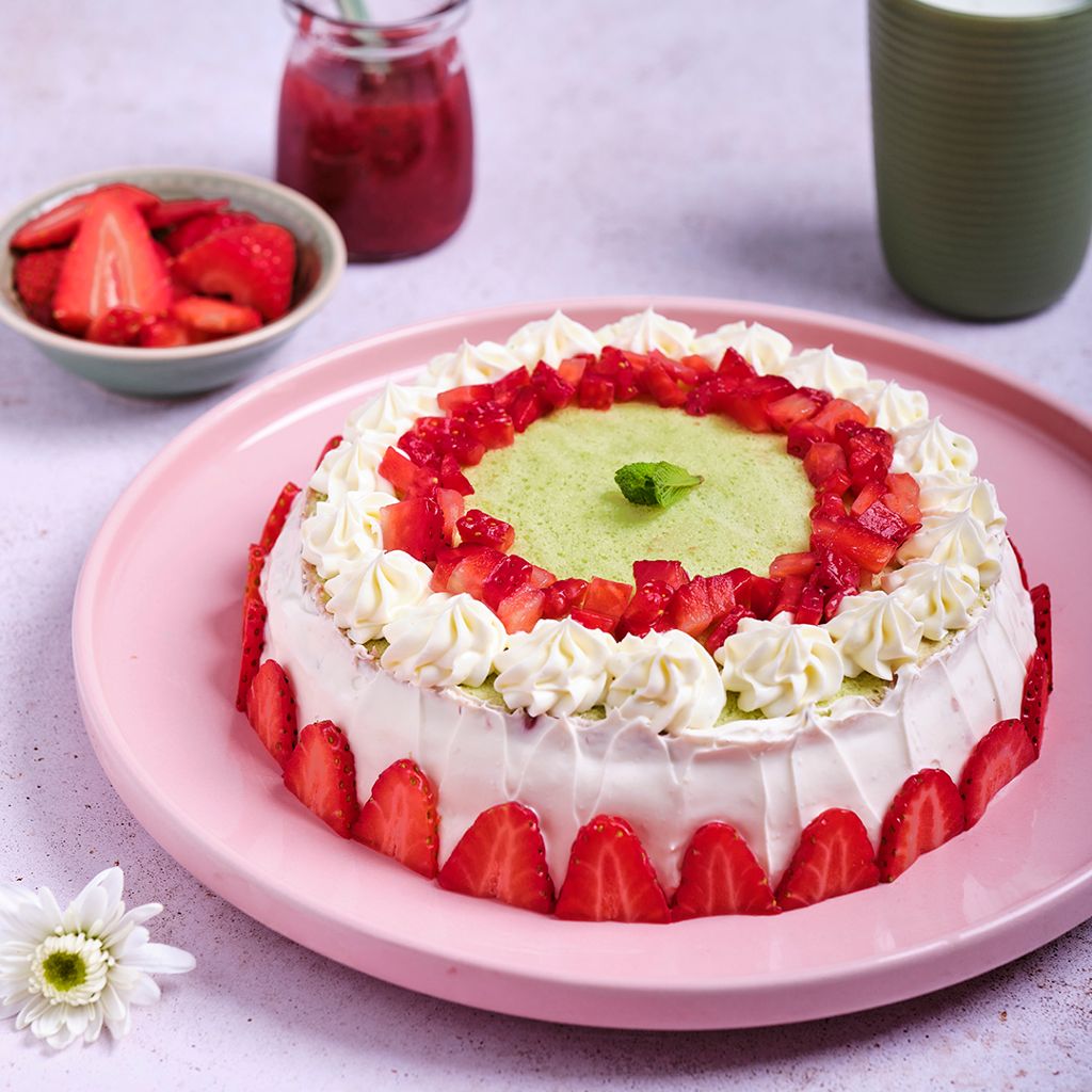A round cake with a green base, white filling and fresh strawberries. Served on a pink plate with a bowl of strawberries alongside.