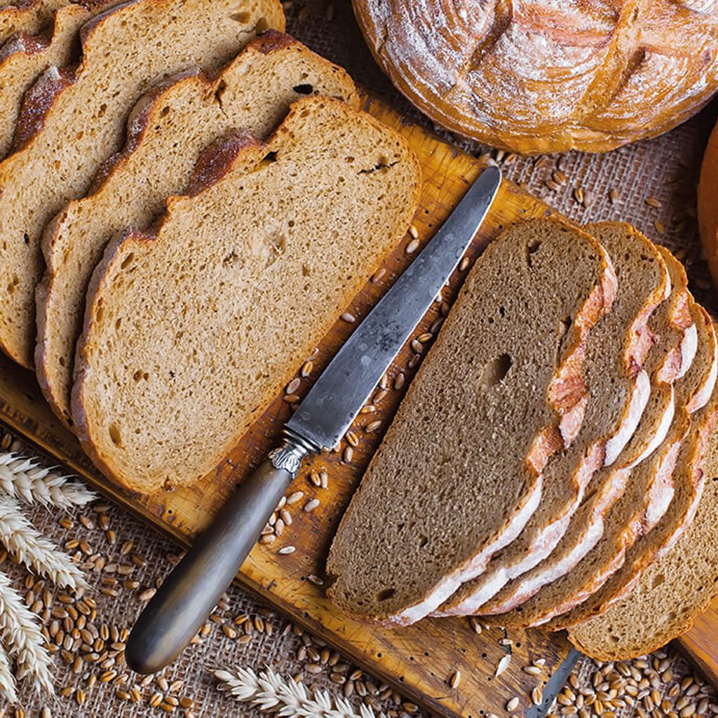 Slices of wholemeal bread arranged on a chopping board with a vintage knife, surrounded by wheat grains and ears of corn.