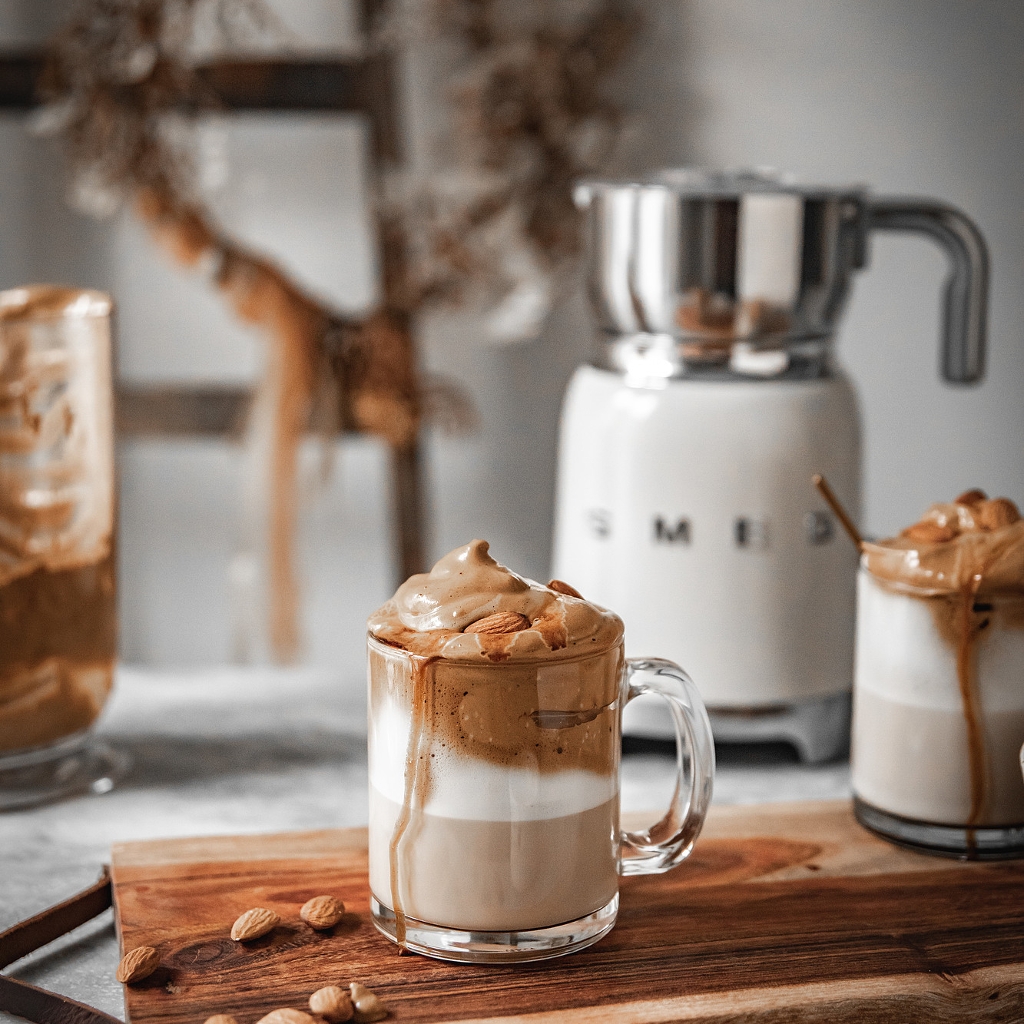 A glass filled with layers of milk and coffee, topped with a thick caramel foam and whole almonds.