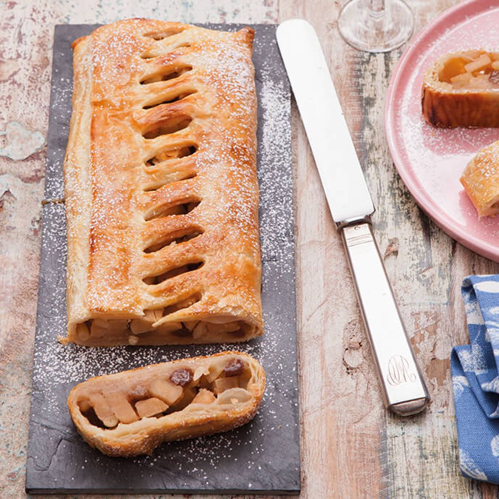 Baked apple strudel dusted with icing sugar on a dark tray, with a slice in the foreground; next to it are knives, a pink plate with more slices, and a decorated blue napkin.