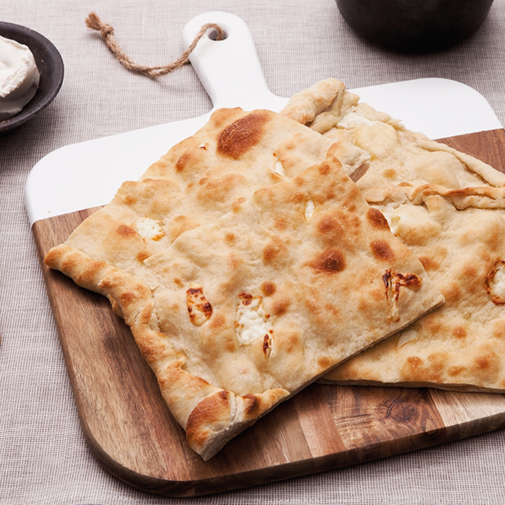 A thin focaccia, baked and topped with cheese, served in slices on a wooden chopping board; alongside it, a bowl of fresh cheese and a knife.
