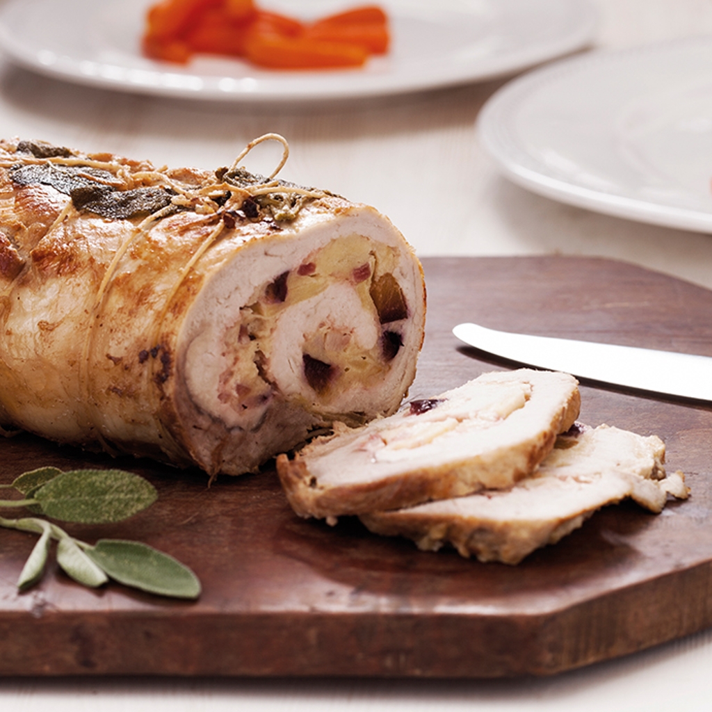 Sliced rolled roast on a chopping board with a knife and sage. In the background, white plates with baby carrots.