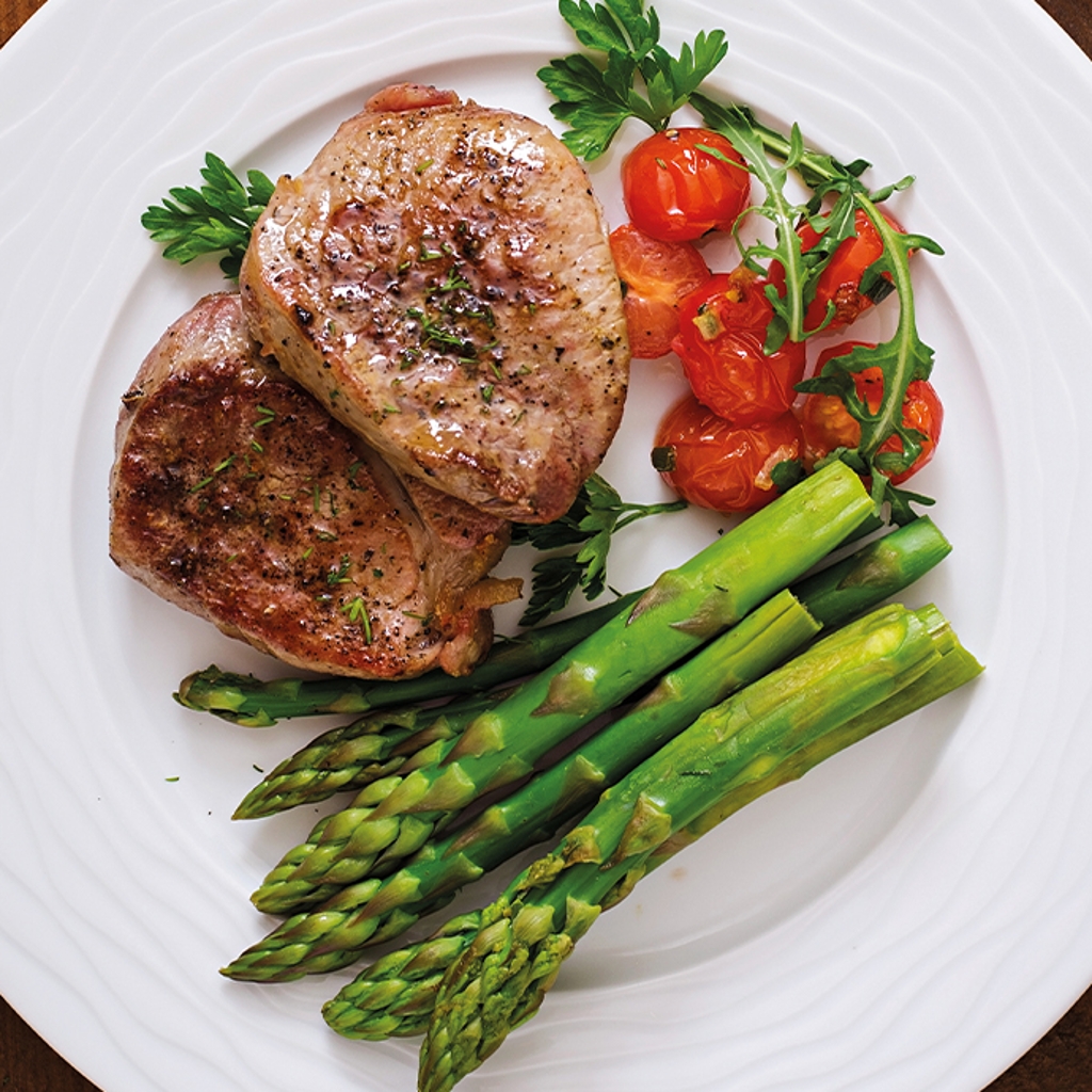 Two fillet medallions with green asparagus and roasted cherry tomatoes, served on a decorated white plate. View from above.