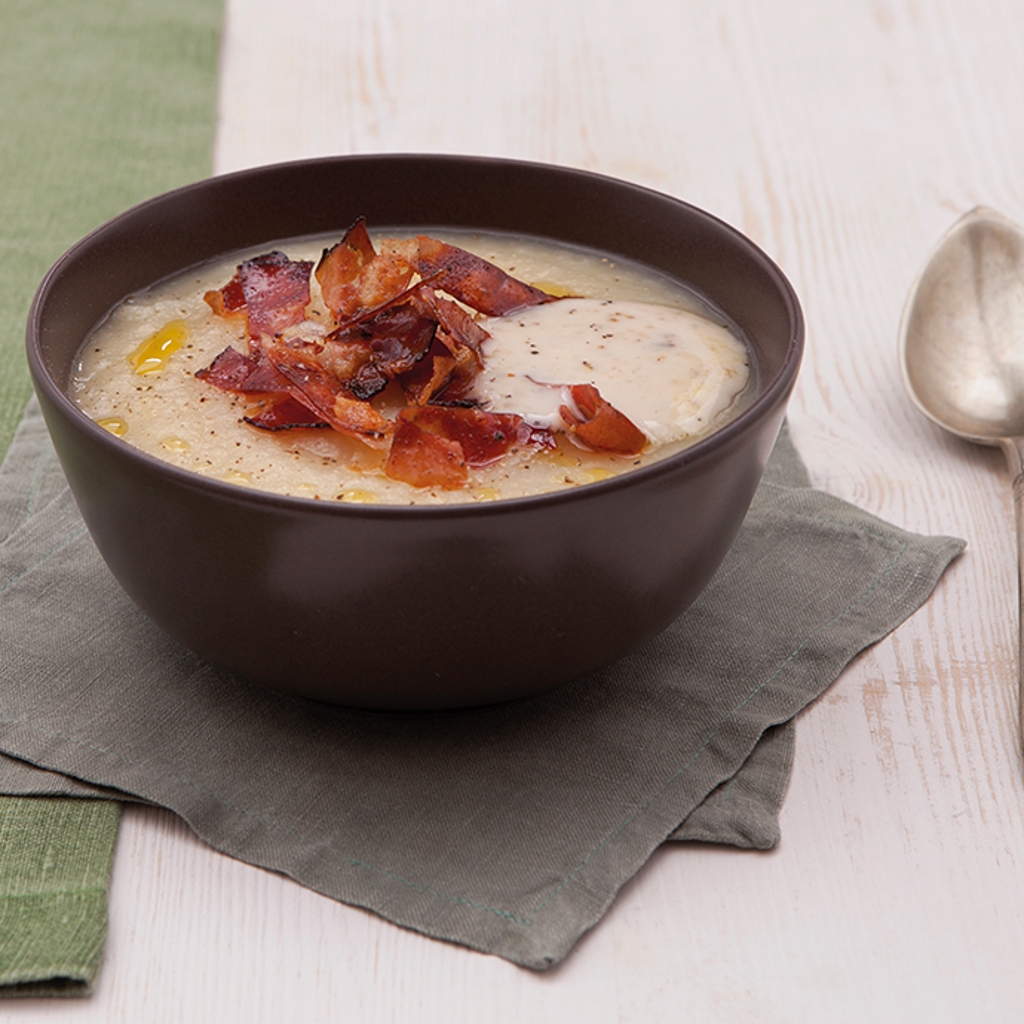 A dark bowl containing a light velouté, crispy speck and truffle cream, served with a spoon on a grey napkin.