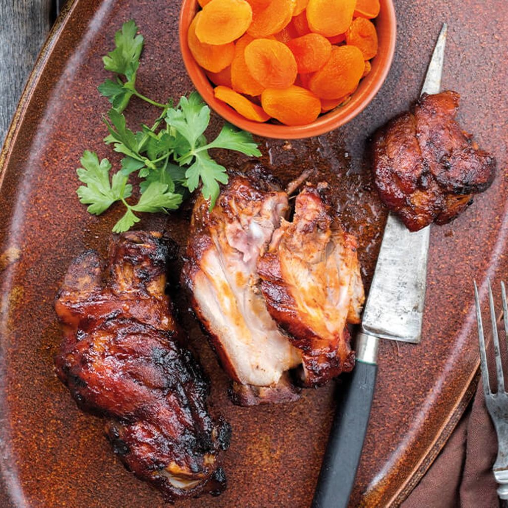 Roast pork shank on a terracotta platter with a bowl of cooked carrots, a knife, a fork and parsley.