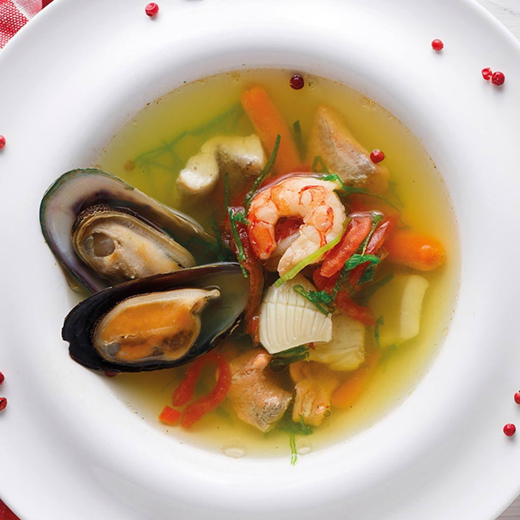 A deep plate of fish soup with mussels, prawns and vegetables in a clear broth, served on a red-and-white chequered tablecloth.