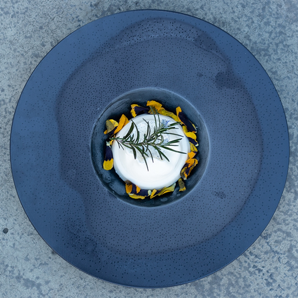A scoop of white ice cream garnished with a sprig of rosemary and yellow petals, placed in the centre of a large, matt black plate.