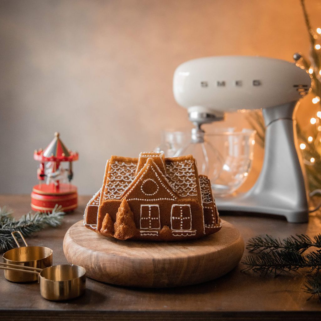 A cake shaped like a little house, decorated with white icing, on a wooden chopping board, with a white Smeg mixer in the background.