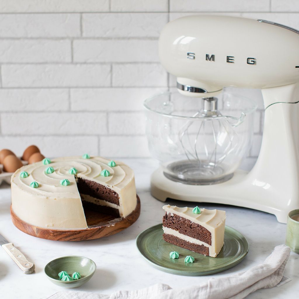 A two-layer chocolate cake with pale frosting and green piping, next to a cream Smeg stand mixer with a glass bowl.