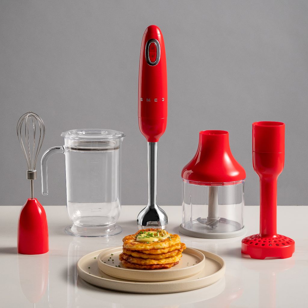 A white Smeg hand mixer next to a plate of potato pancakes, a bowl of sour cream, and a bowl of raw potatoes.