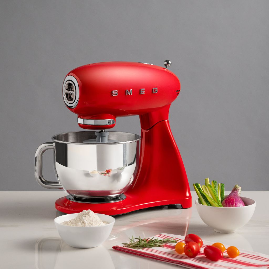 Red Smeg retro stand mixer on a counter with a bowl of flour, herbs, and tomatoes, prepared for baking.