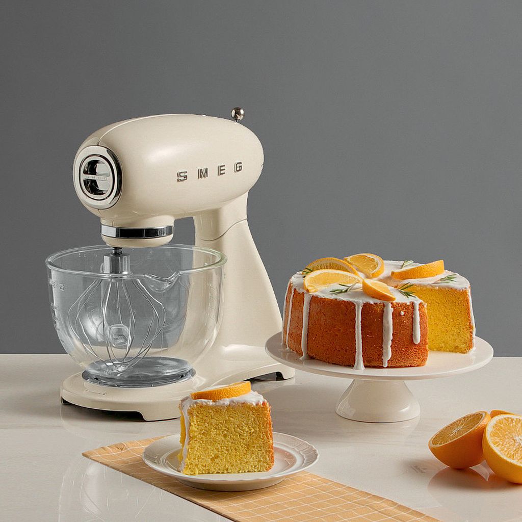 Slices of vibrant orange chiffon cake with white glaze and orange segments, displayed next to a cream Smeg stand mixer with a glass bowl.